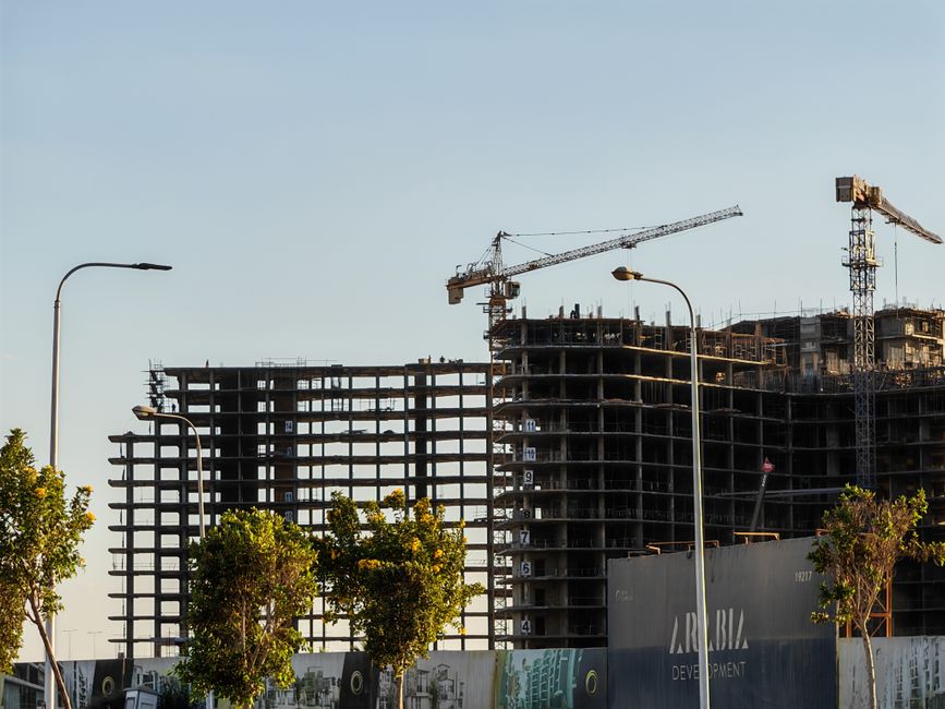 High-rise building under construction with cranes and trees in the forefront.