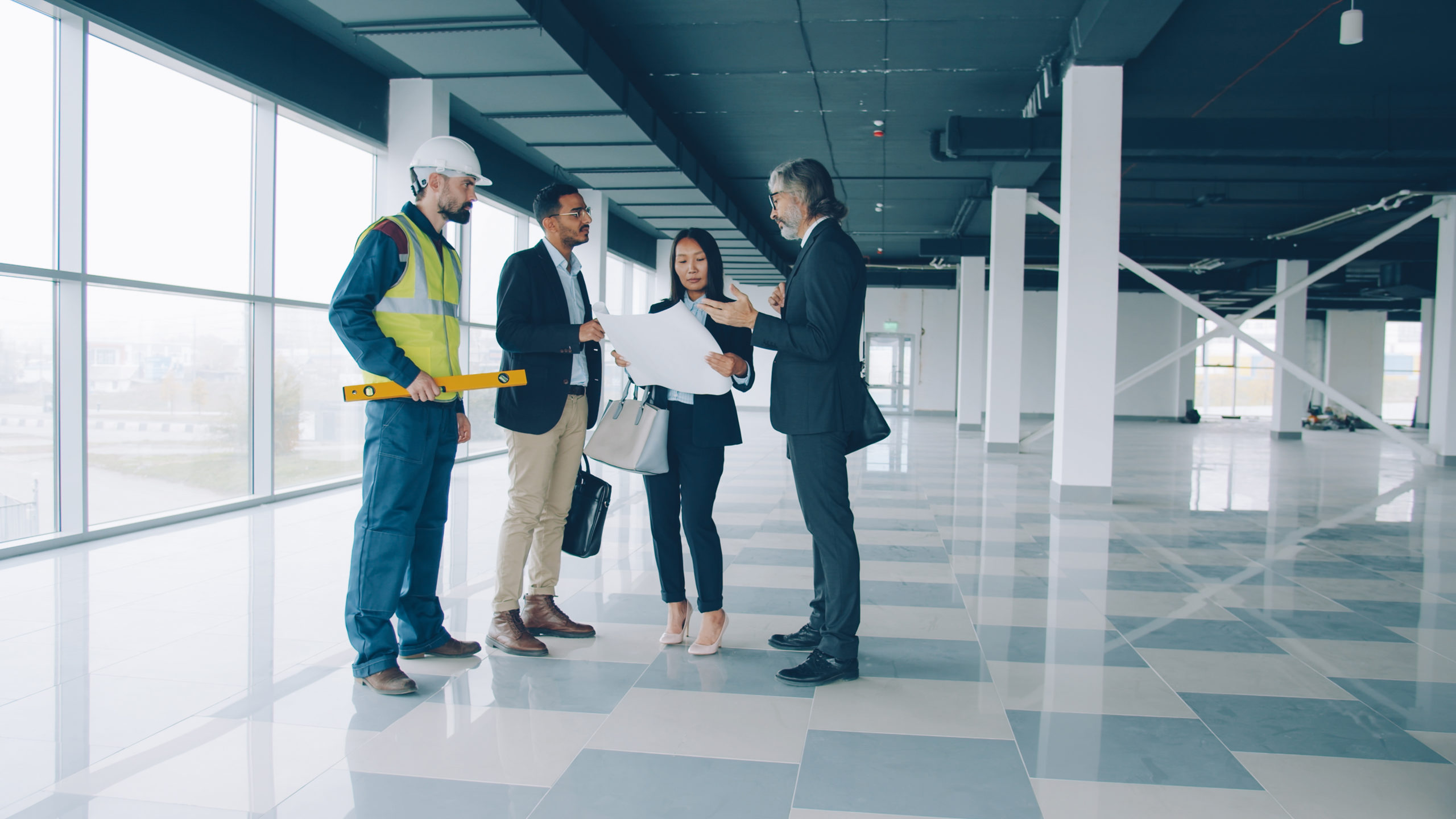 Group of four businesspeople discussing blueprints inside modern commercial office building.