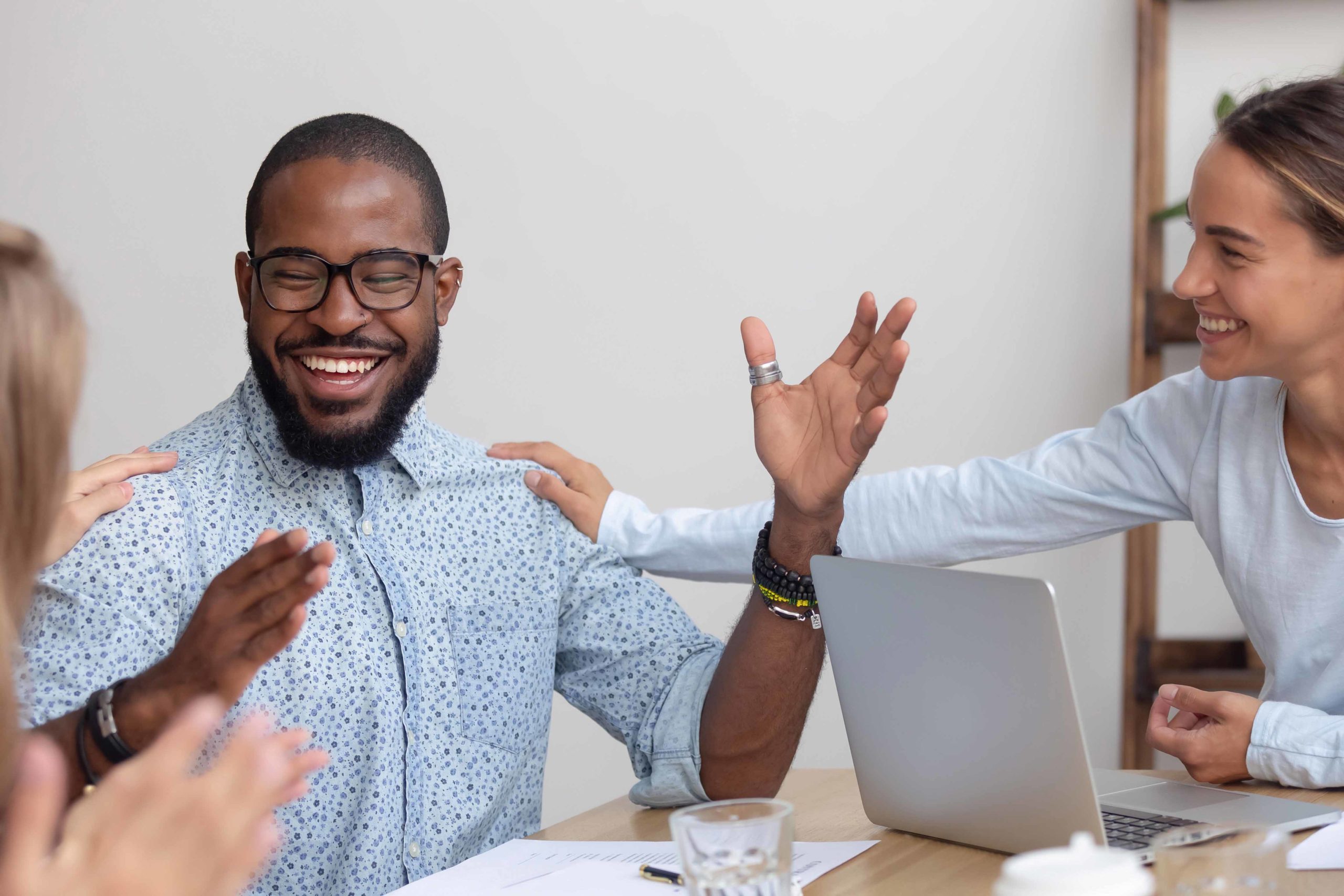 Friendly female caucasian team employees tap african american male colleague on the shoulder supporting cheering mate congratulating happy black coworker with promotion reward, recognition concept