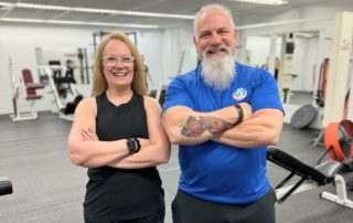 Smiling woman with her personal trainer inside the Prescription Fitness training center gym, both with arms crossed, showing confidence after a fitness session.