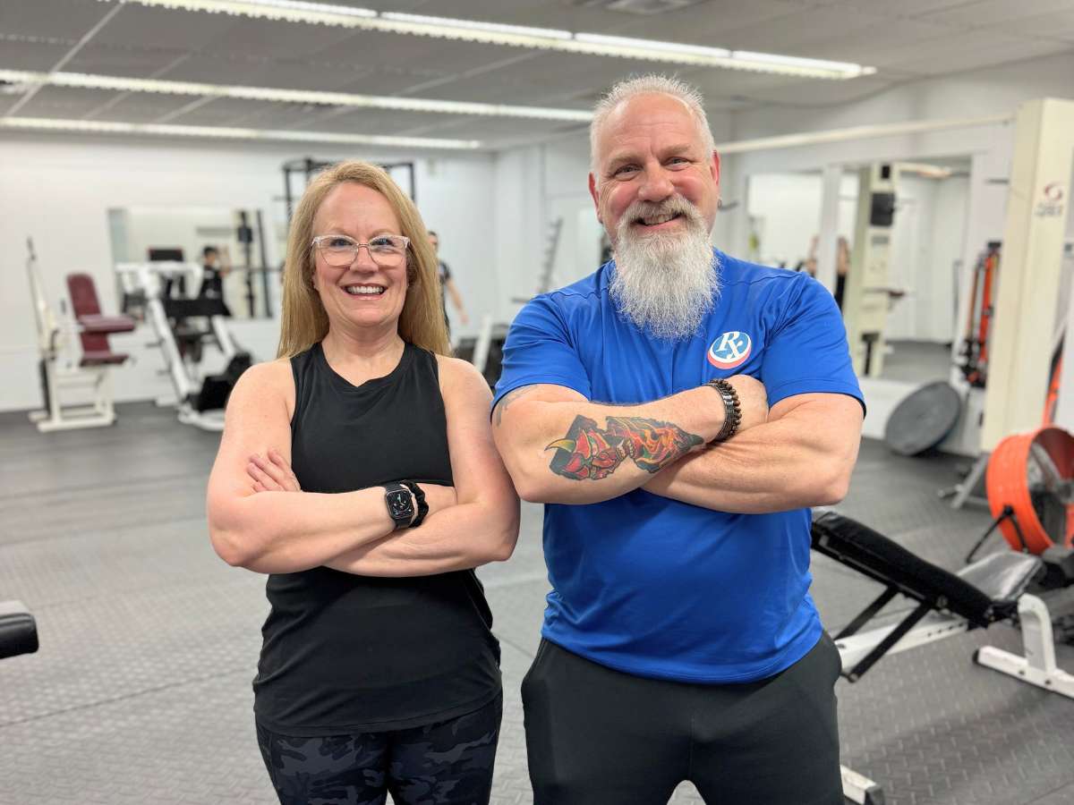 Smiling woman with her personal trainer inside the Prescription Fitness training center gym, both with arms crossed, showing confidence after a fitness session.