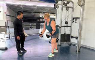 Rosanne Barnhart performing a bent-over cable fly while her trainer observes during a personal training session.