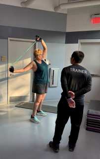Rosanne Barnhart stretches a resistance band overhead while her trainer observes during a workout session.