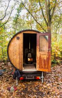Rustic outdoor barrel sauna on a trailer, surrounded by autumn leaves and trees, with a warm fire glowing inside.