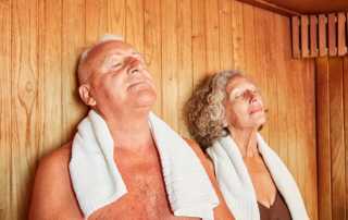 Older man and woman relaxing in a sauna with towels around their necks, eyes closed, enjoying the heat.