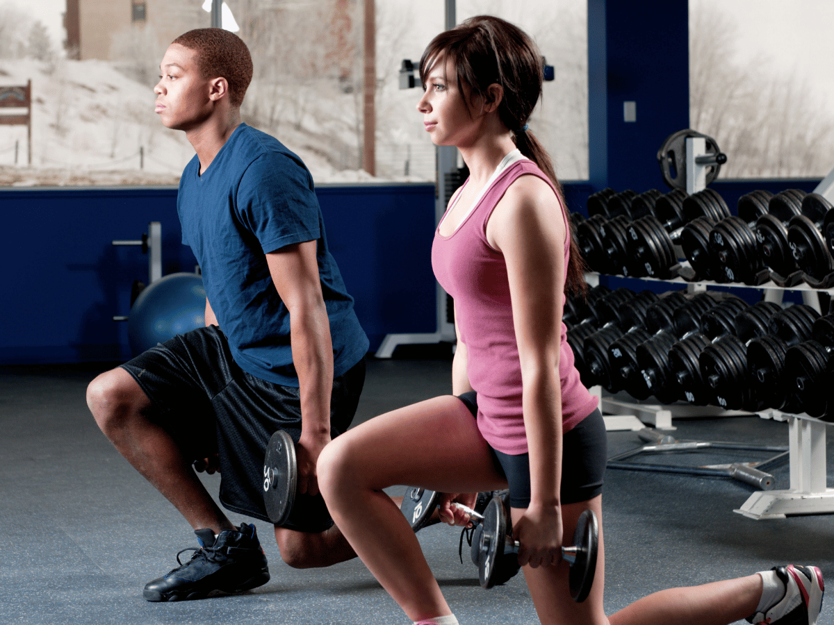 teen strength training program Two people performing walking lunges with dumbbells in a gym, focusing on proper form and balance.