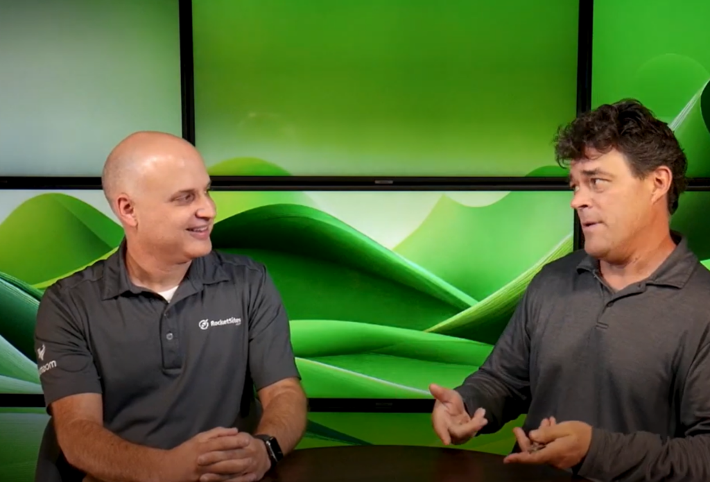 Two men sitting at a table having a conversation in front of a green abstract background displayed on large screens.