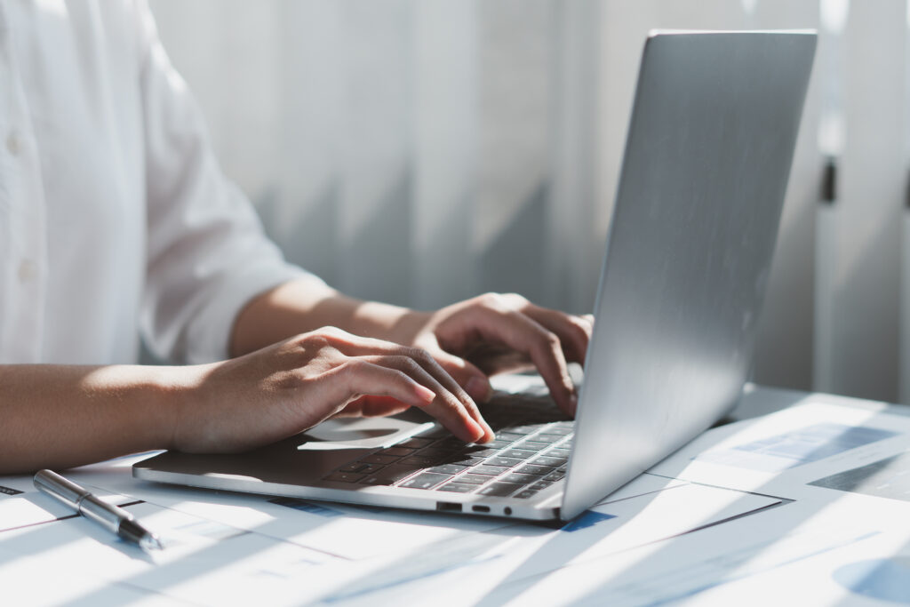 Close-up of a person typing on a laptop keyboard with sunlight streaming through window blinds in the background.