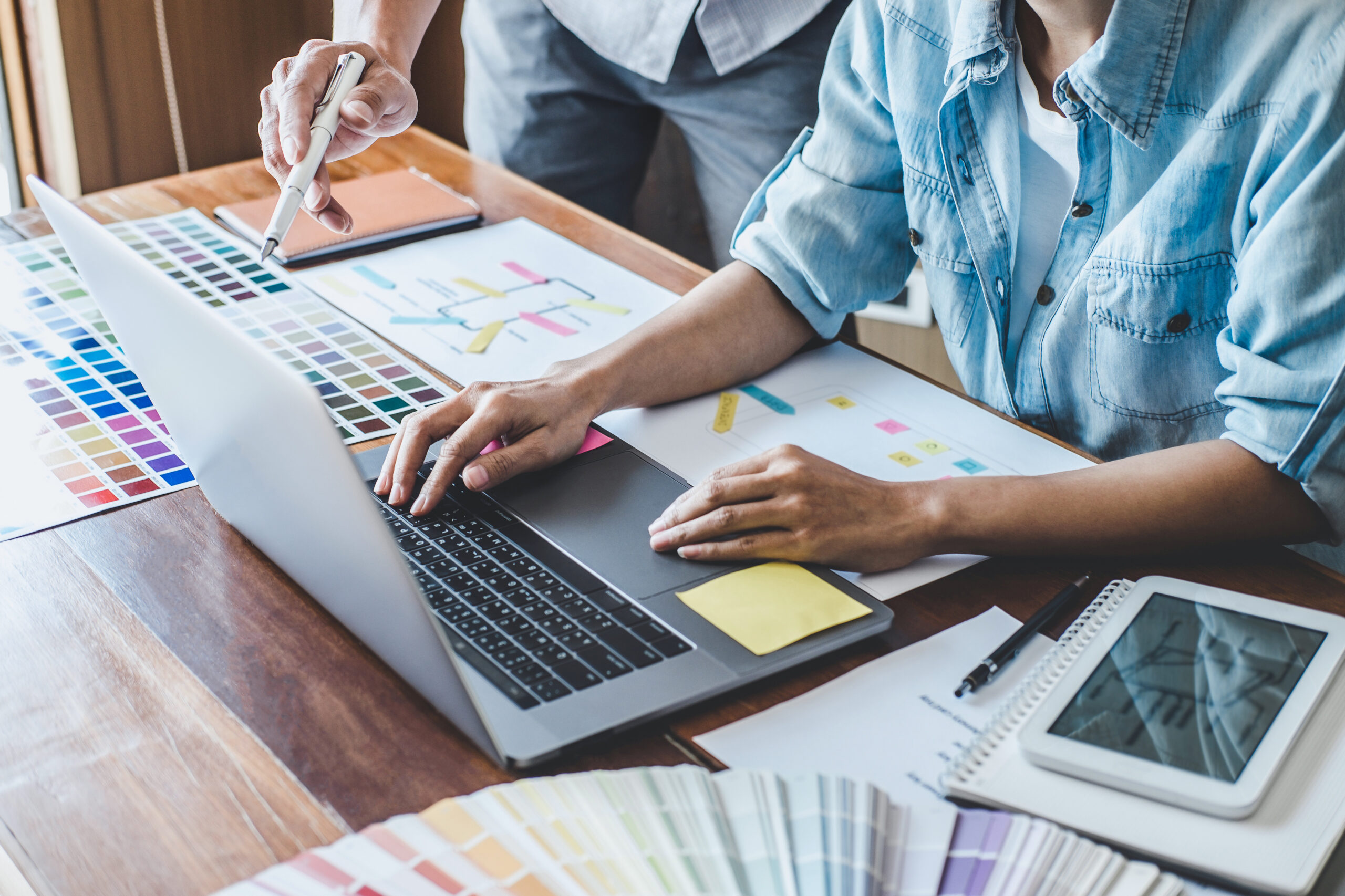Close-up of a designer's hands working on a laptop surrounded by color swatches, wireframes, and printed layout designs during a creative planning session.