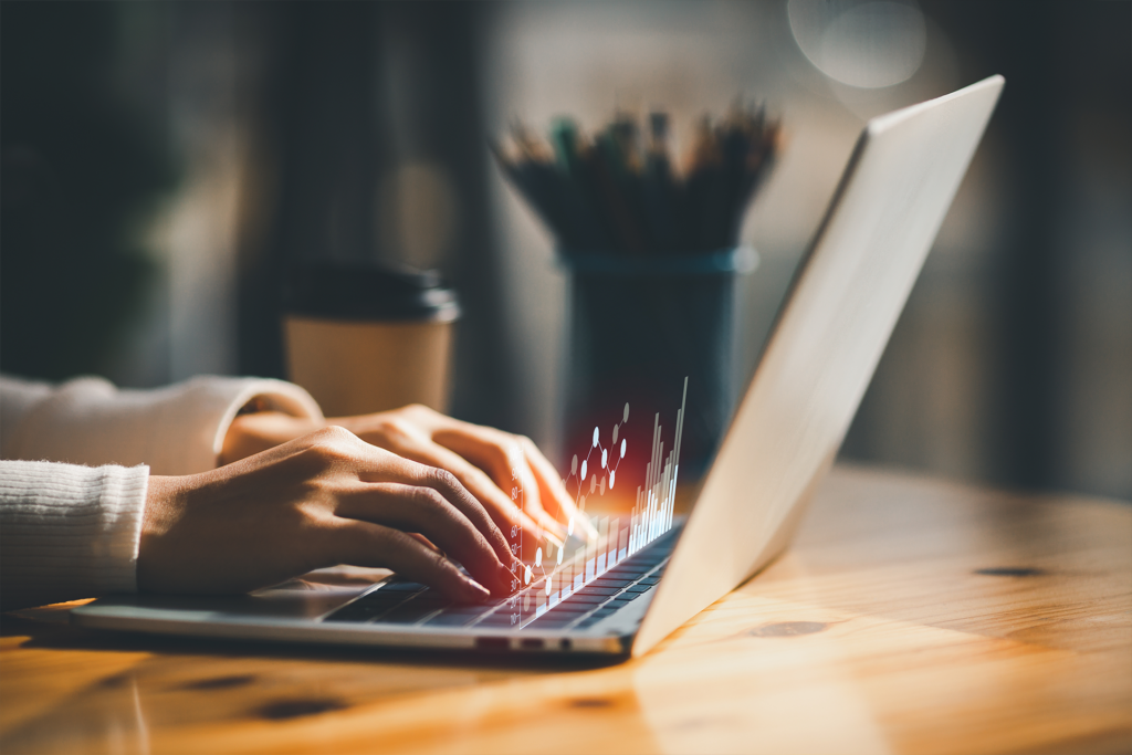 Close-up of hands typing on a laptop with digital data graphs overlaid, a coffee cup and pencil holder blurred in the background on a wooden desk