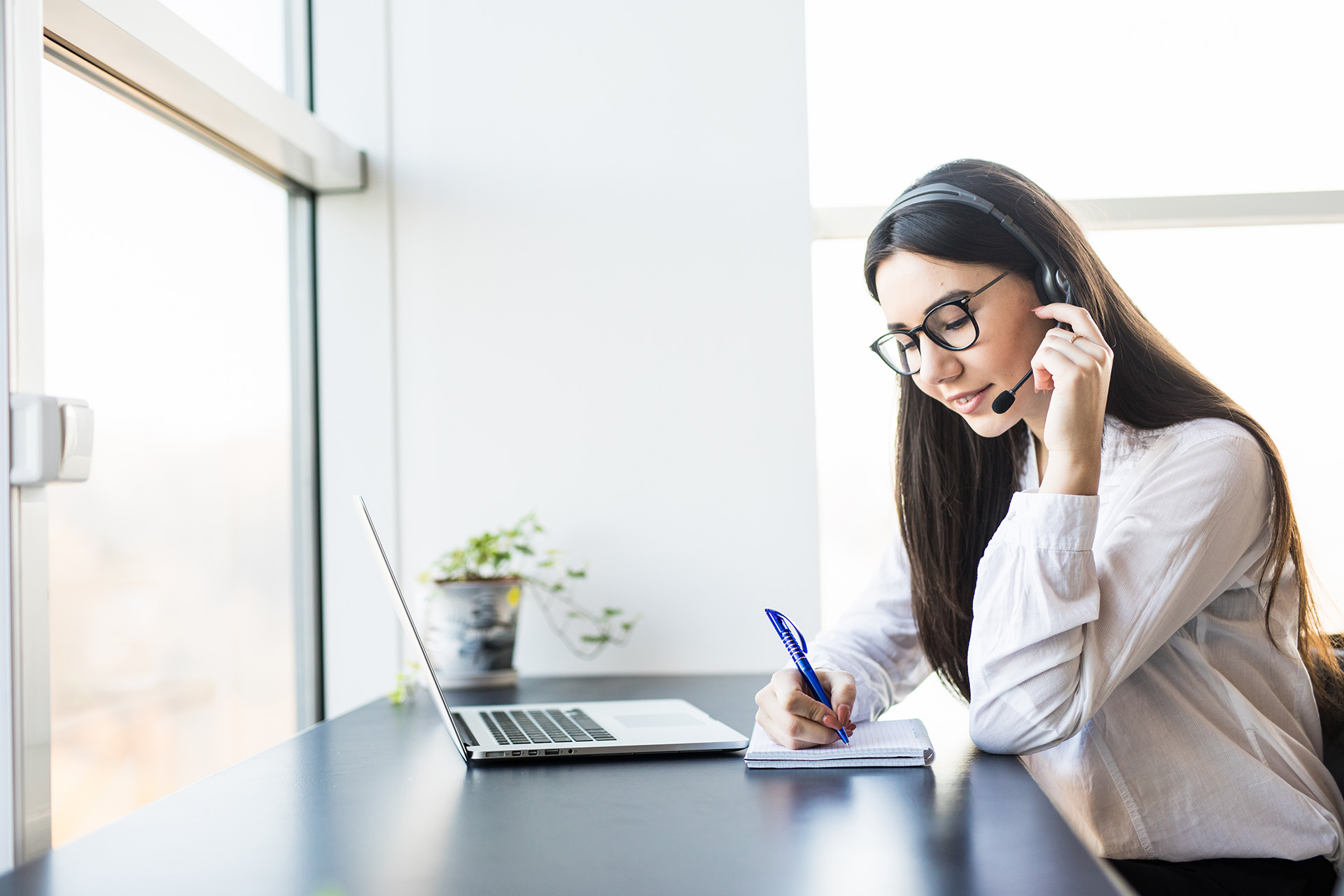 Woman wearing a headset and glasses taking notes in a notebook while attending a video call on her laptop in a bright office space.